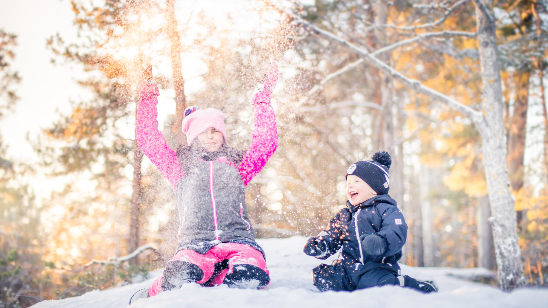 Two small children are throwing snow into the air and laughing in front of several trees.