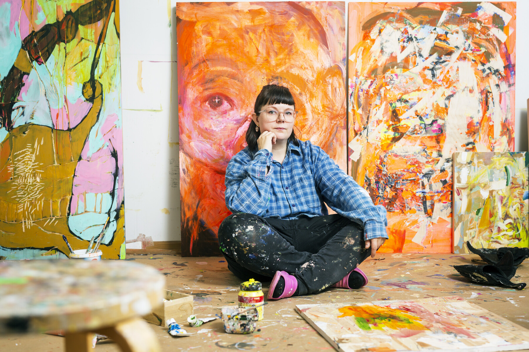 Eine Frau in Arbeitskleidung sitzt auf dem Boden eines Ateliers vor mehreren großen Gemälden, die an die Wand gelehnt sind.
