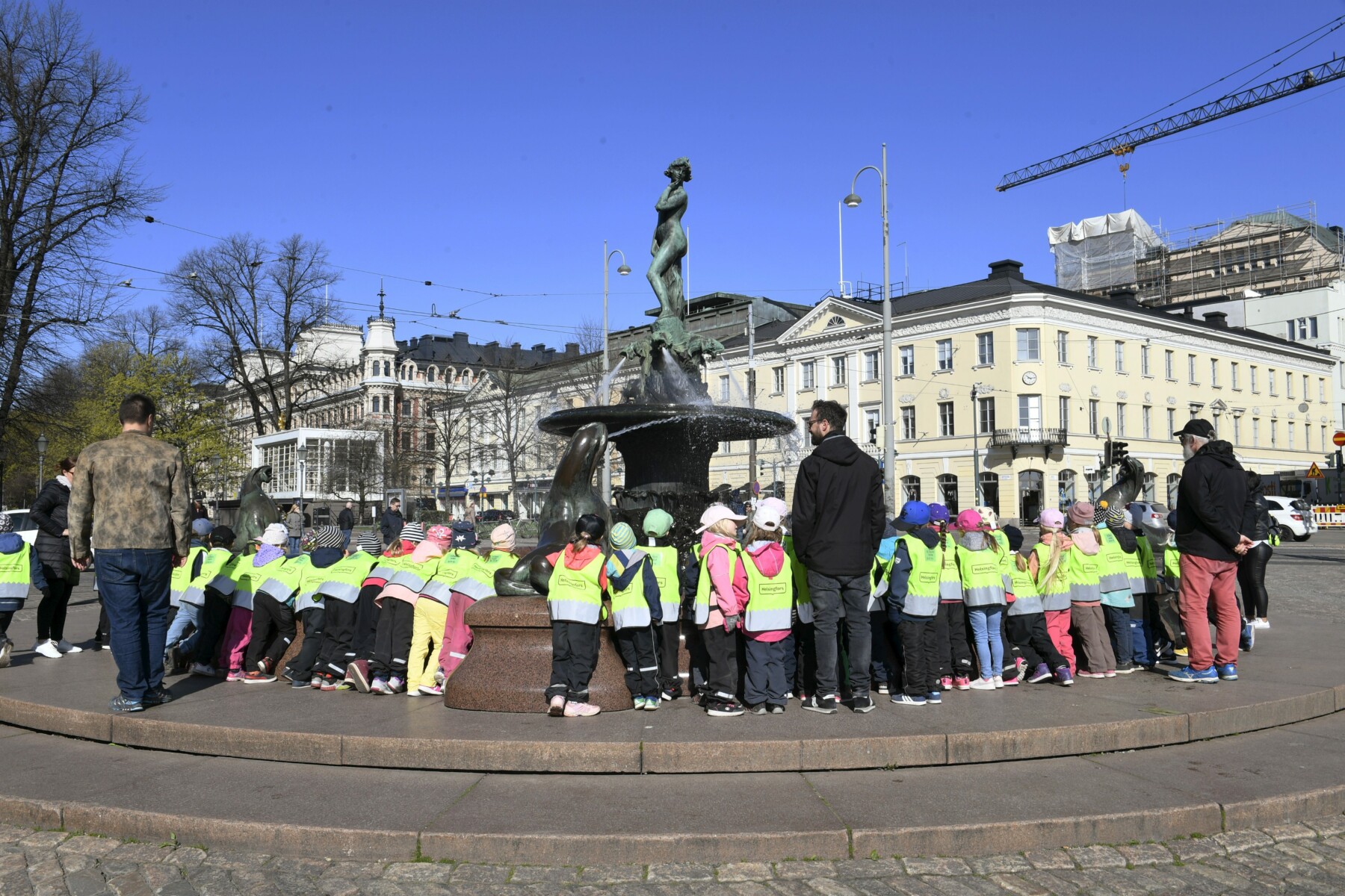 On a plaza with buildings in the background, several dozen children and teachers are gathered around a metal statue on a tall pedestal.