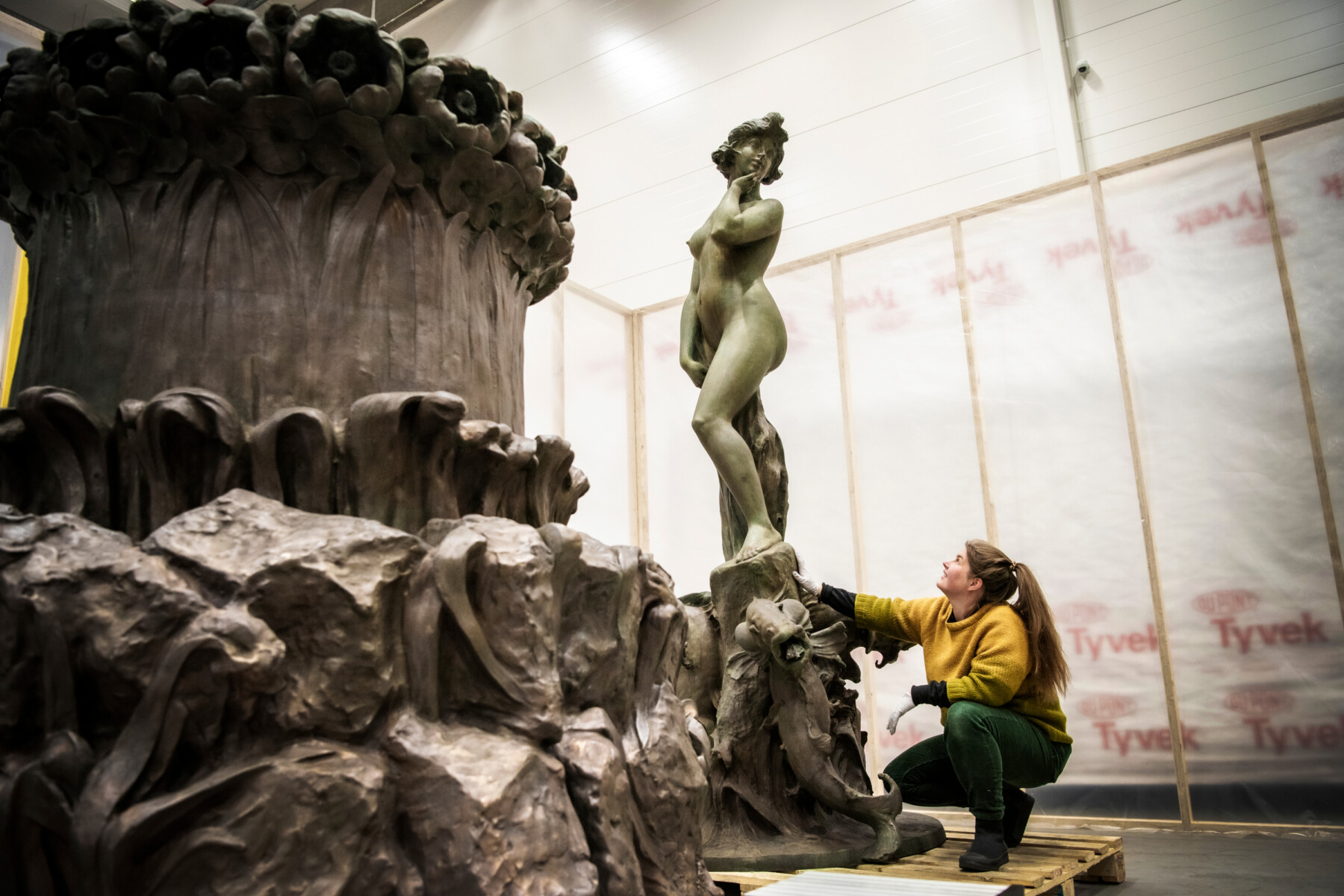 A woman kneels to look at the base of a statue in a workshop in front of a metal statue of a female figure.
