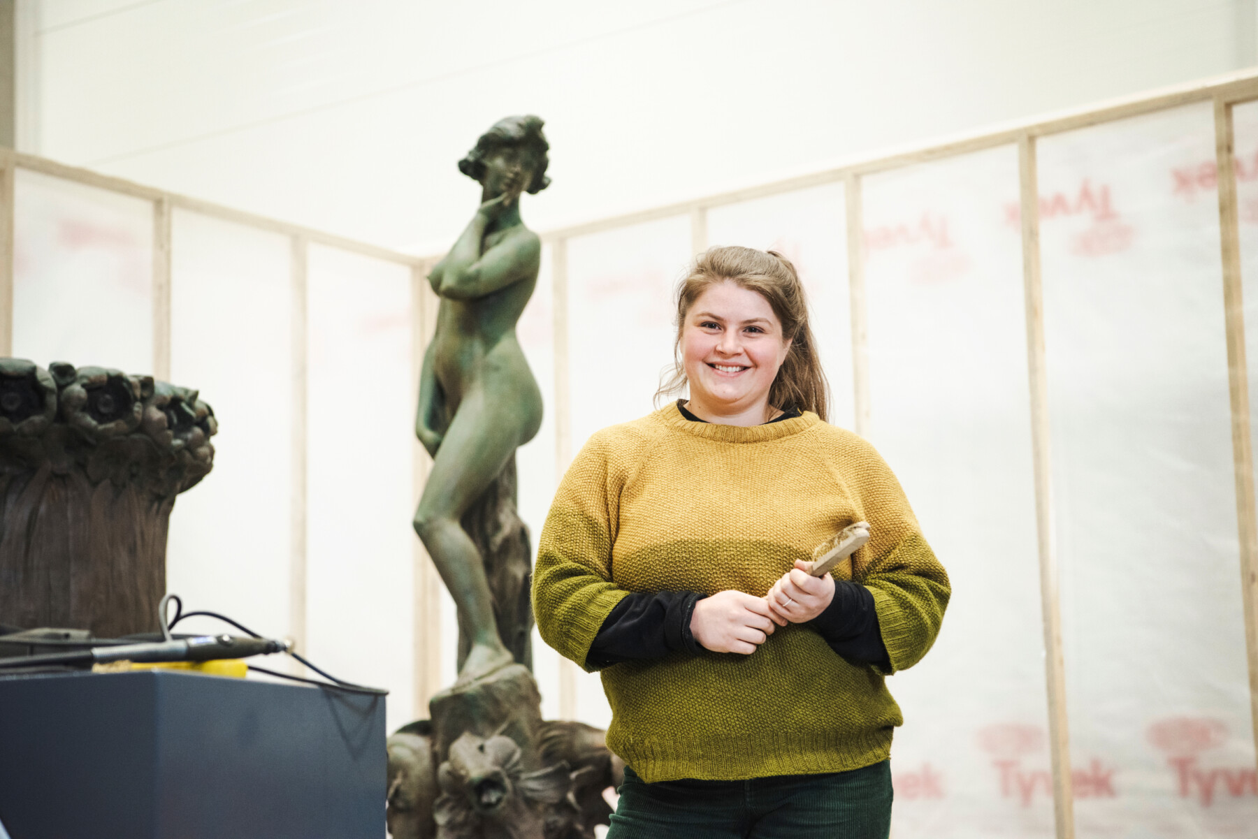 Uma mulher com um pincel nas mãos está em uma oficina em frente a uma estátua de metal de uma figura feminina.