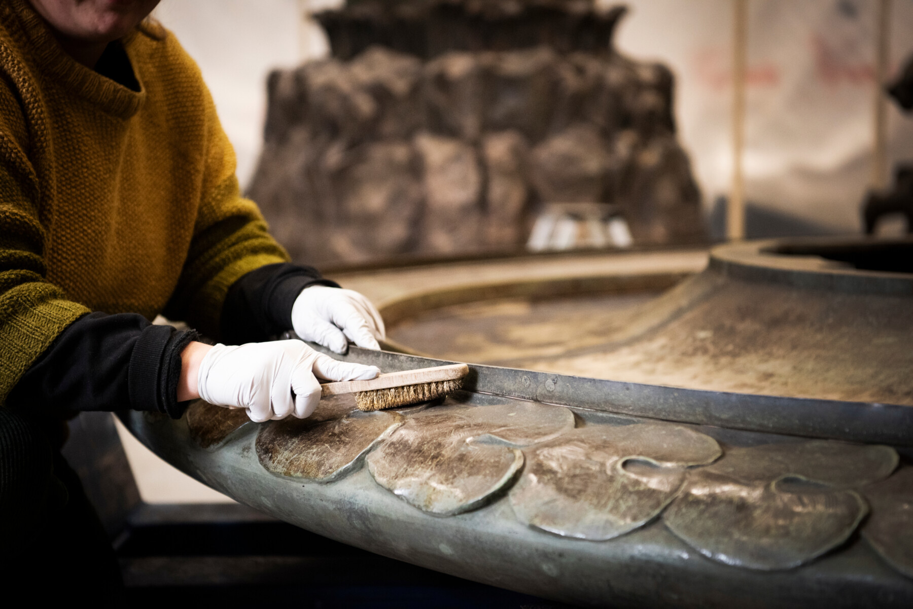 Un gros plan présente deux mains gantées en train d’utiliser une brosse pour nettoyer les motifs en forme de feuilles présents autour d'une partie de la base d'une sculpture en métal. 
