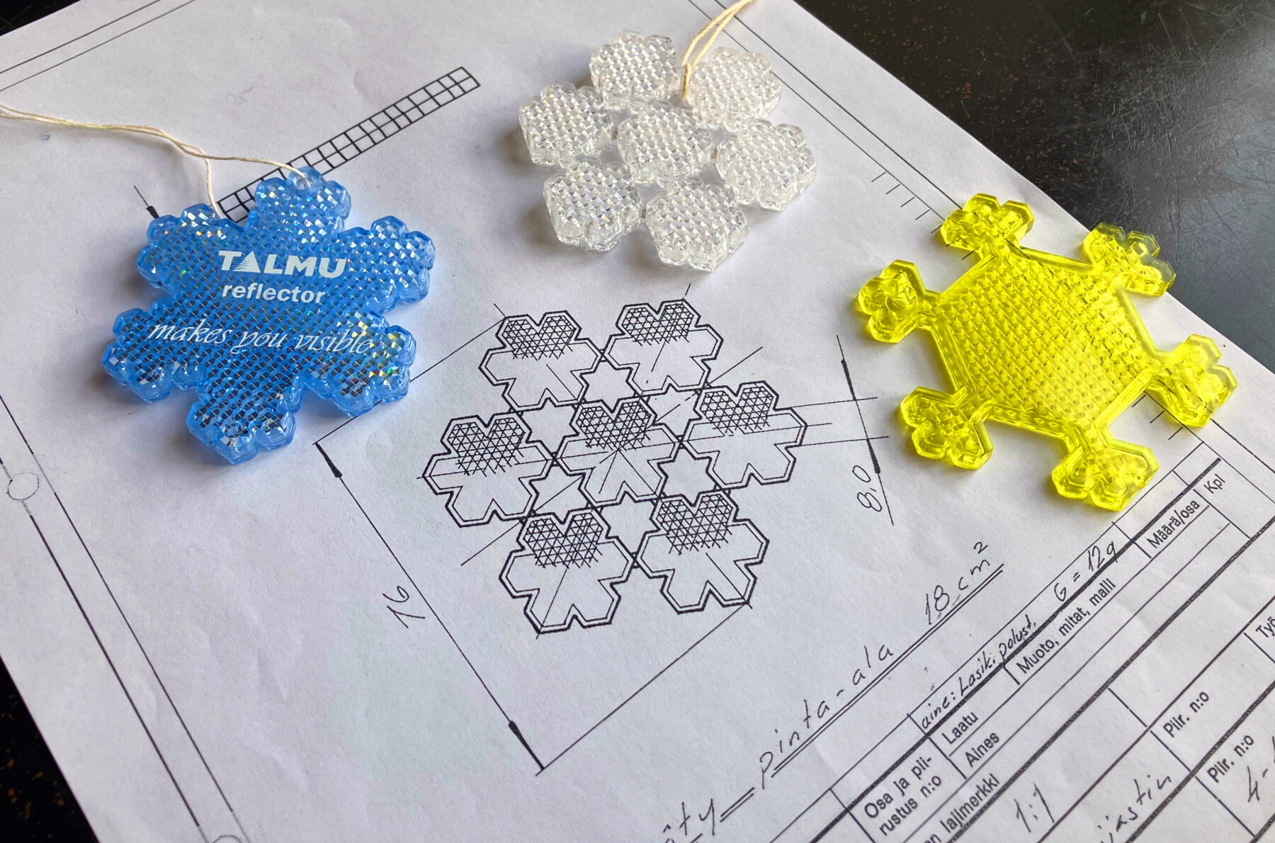 Tres piezas planas de plástico de seis puntas y cada una de distinto color, dispuestas sobre un papel que muestra un dibujo técnico, también en forma de estrella.