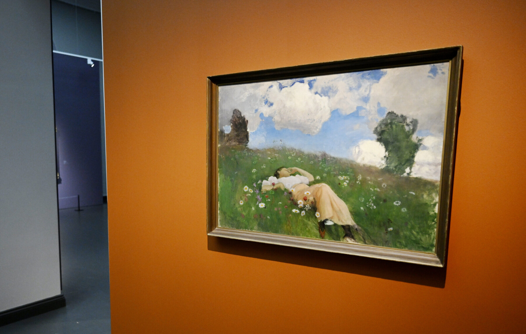 A painting hanging in a gallery shows a woman lying in a field of flowers under a blue sky with scattered clouds.