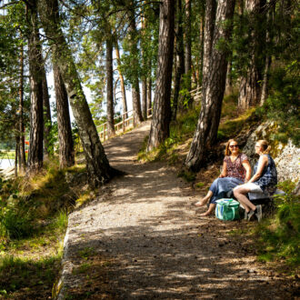 Deux femmes en tenue estivale sont assises sur un banc dans une forêt au bord d'un sentier qui suit la rive d'un lac.