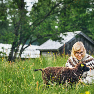 A woman is kneeling to pet a lamb in a green meadow in front of several old wooden buildings.