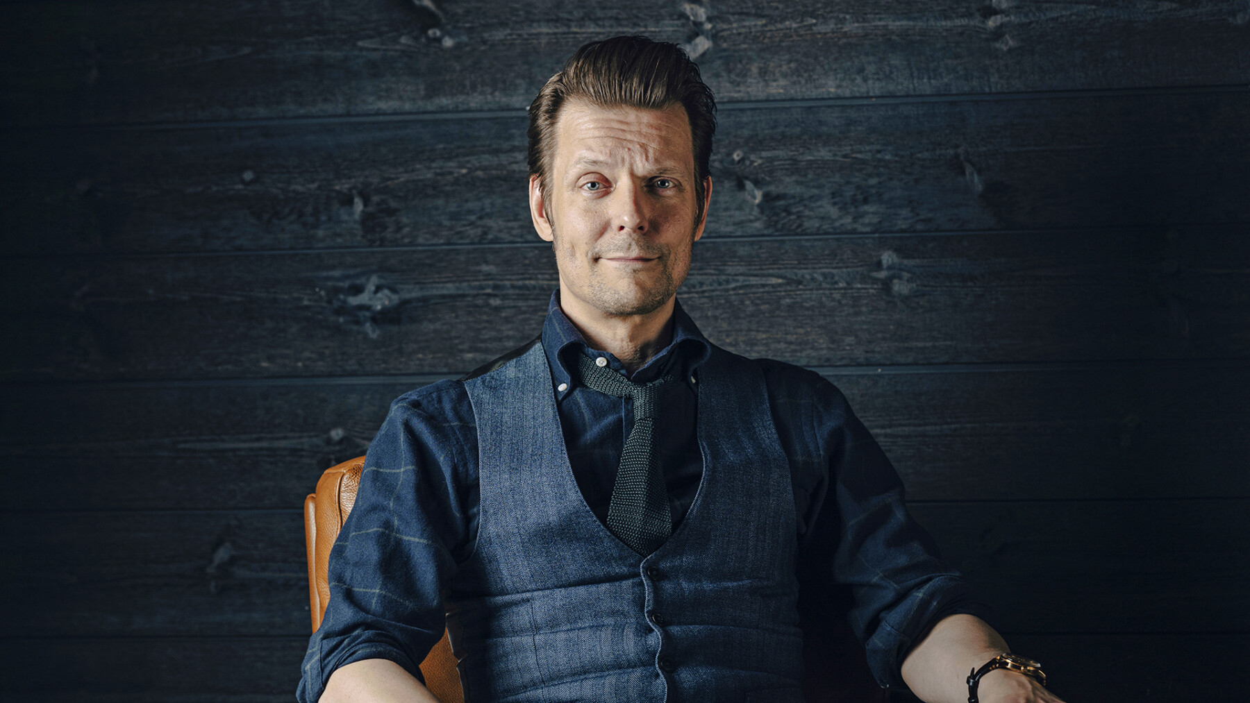 A man in a dark blue shirt and vest is sitting in a chair, looking into the camera.