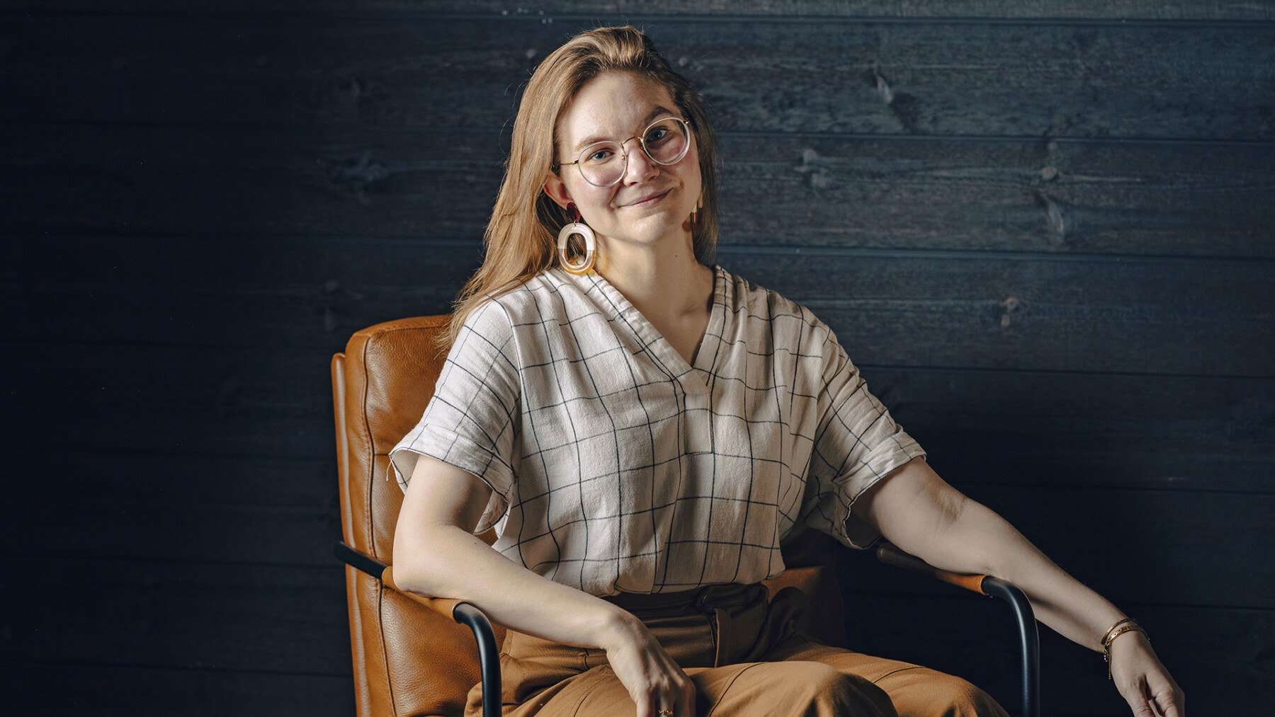 A woman with round glasses and round earrings is sitting in a chair, looking into the camera.