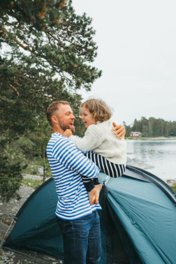 A man holding a child in his arms with a small tent and a lake in the background.