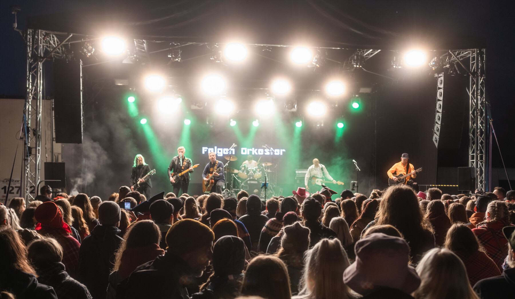 At an outdoor night concert, a crowd watches a band consisting of five guitarists and a drummer.