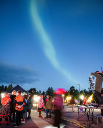 A green band of light is visible in the twilight over the festival, where a bunch of people are standing and talking.