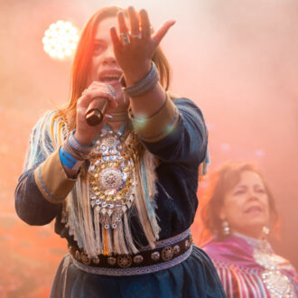 Duas mulheres em vestidos ornamentados estão cantando em microfones em um palco sob holofotes.