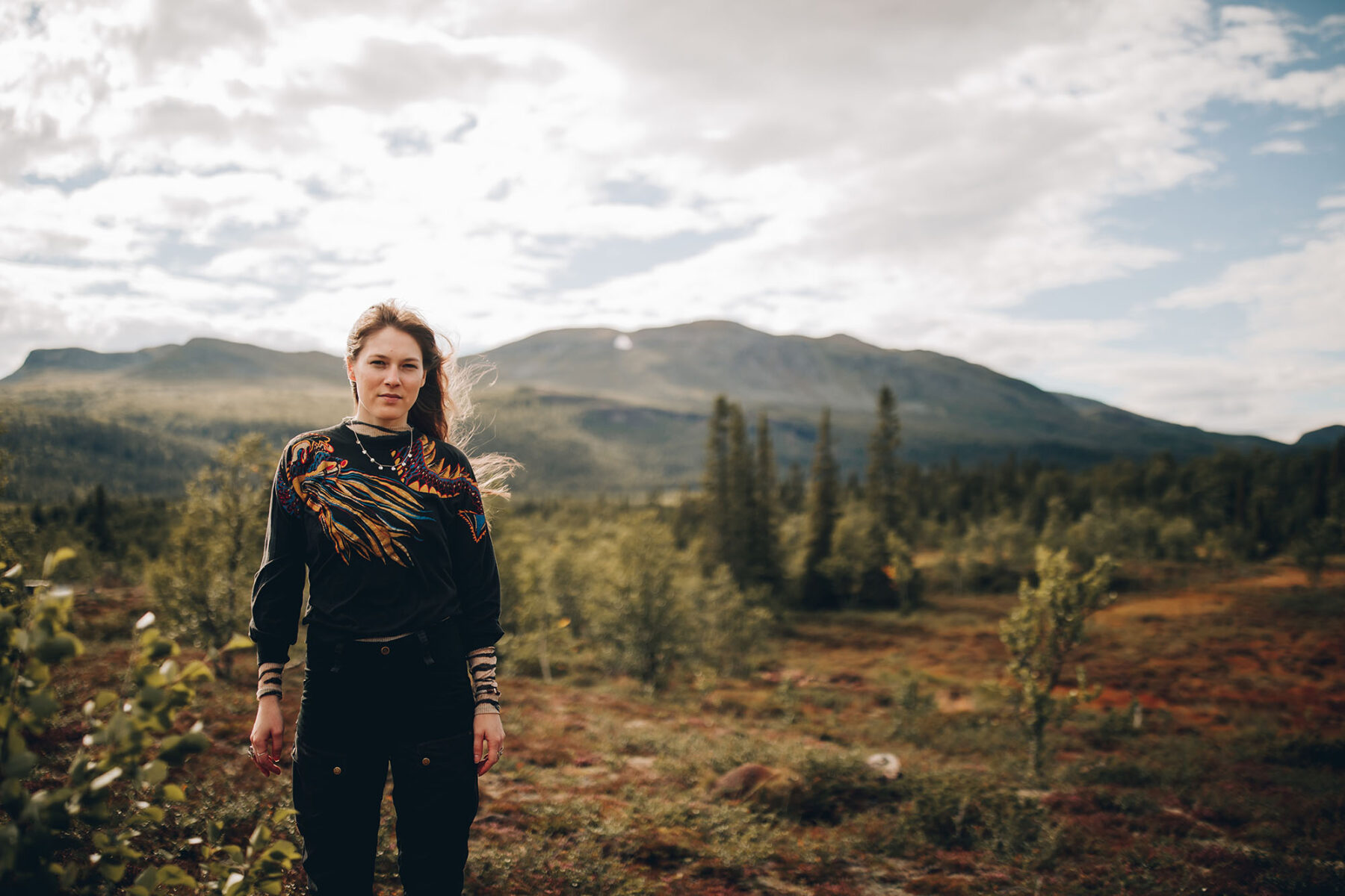 Une femme pose au milieu d’un paysage où des montagnes se profilent en arrière-plan. 