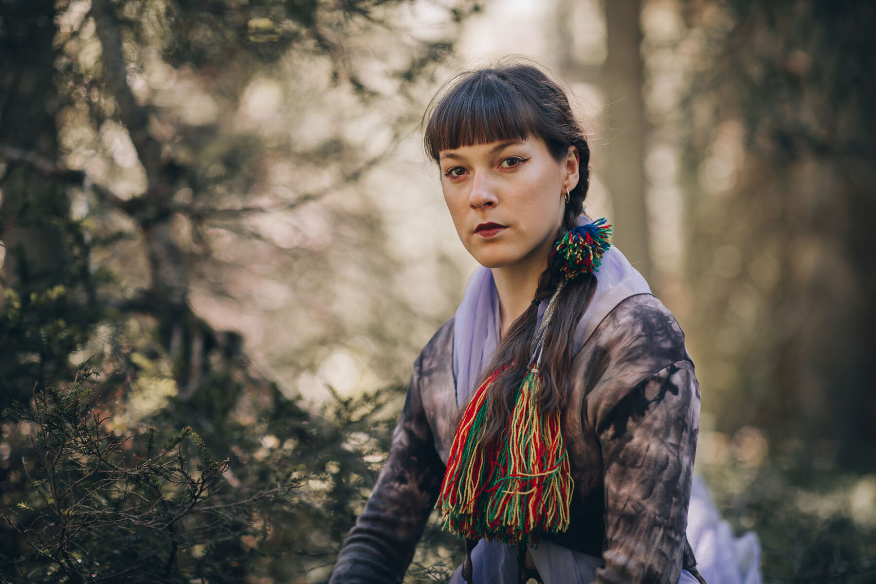 A woman looks into the camera with a serious expression, with a forest in the background.