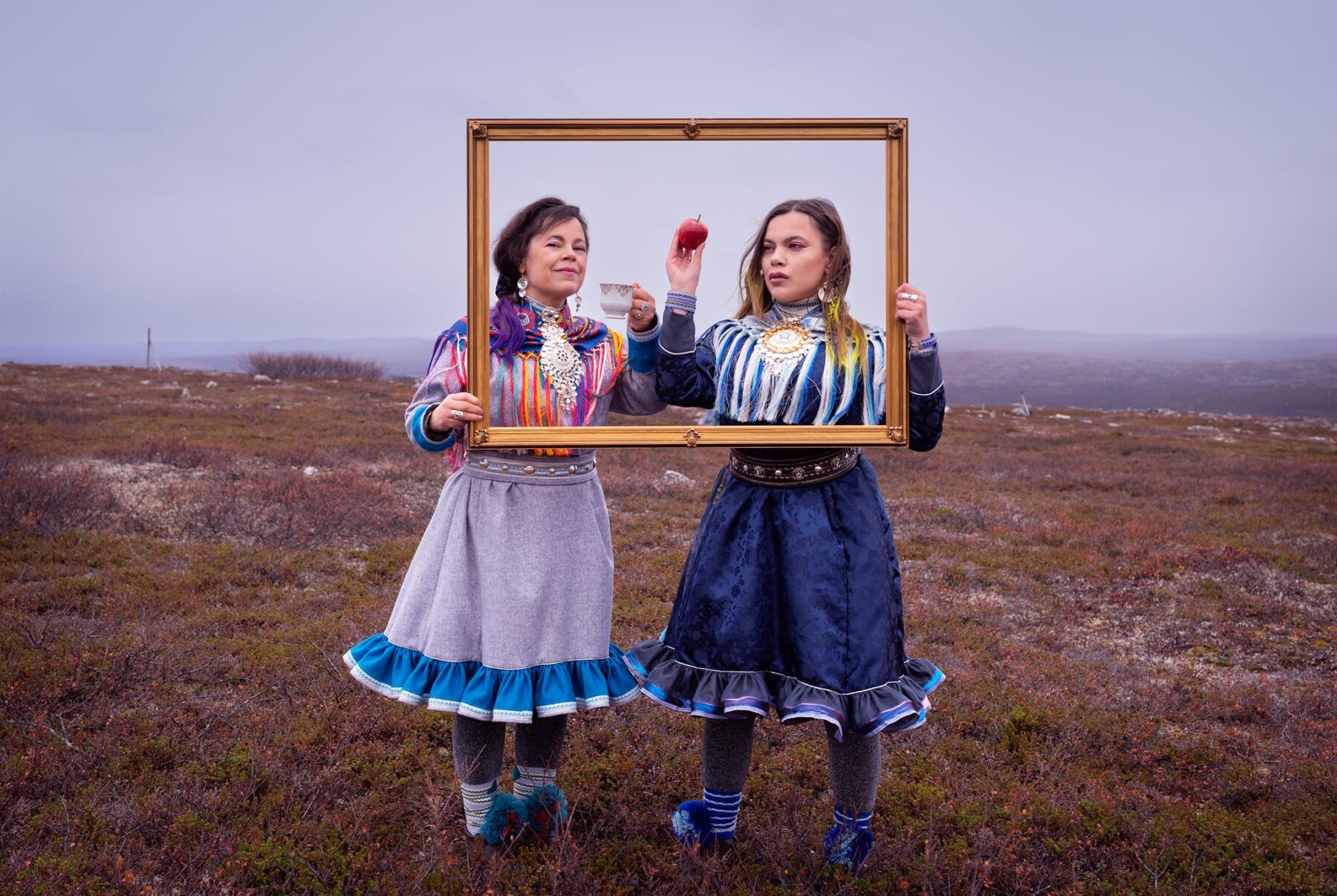 Two women in ornate dresses pose outside with mountains in the background, holding an empty picture frame up in front of their heads and shoulders.