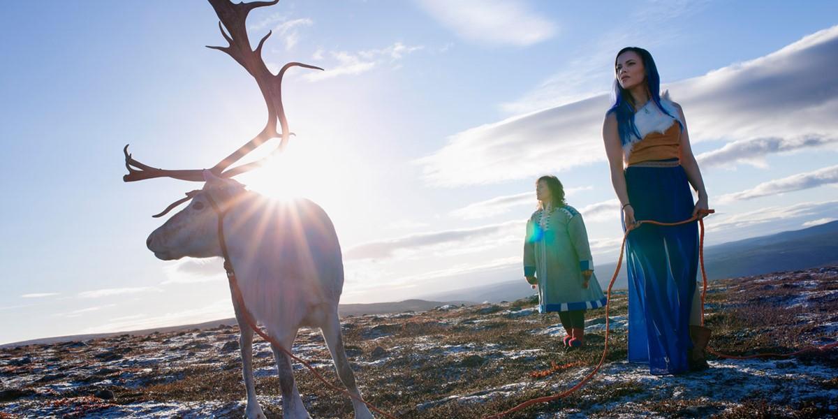 Outside with mountains in the background, two women in ornate dresses pose near a reindeer with large antlers.