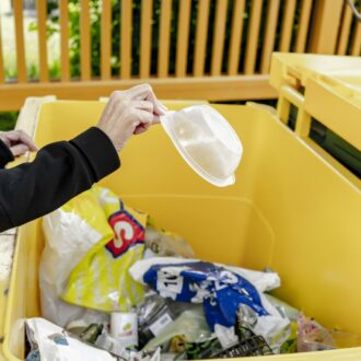 En un punto de recogida de residuos domésticos, una persona tira una bolsa de plástico en un gran contenedor amarillo.