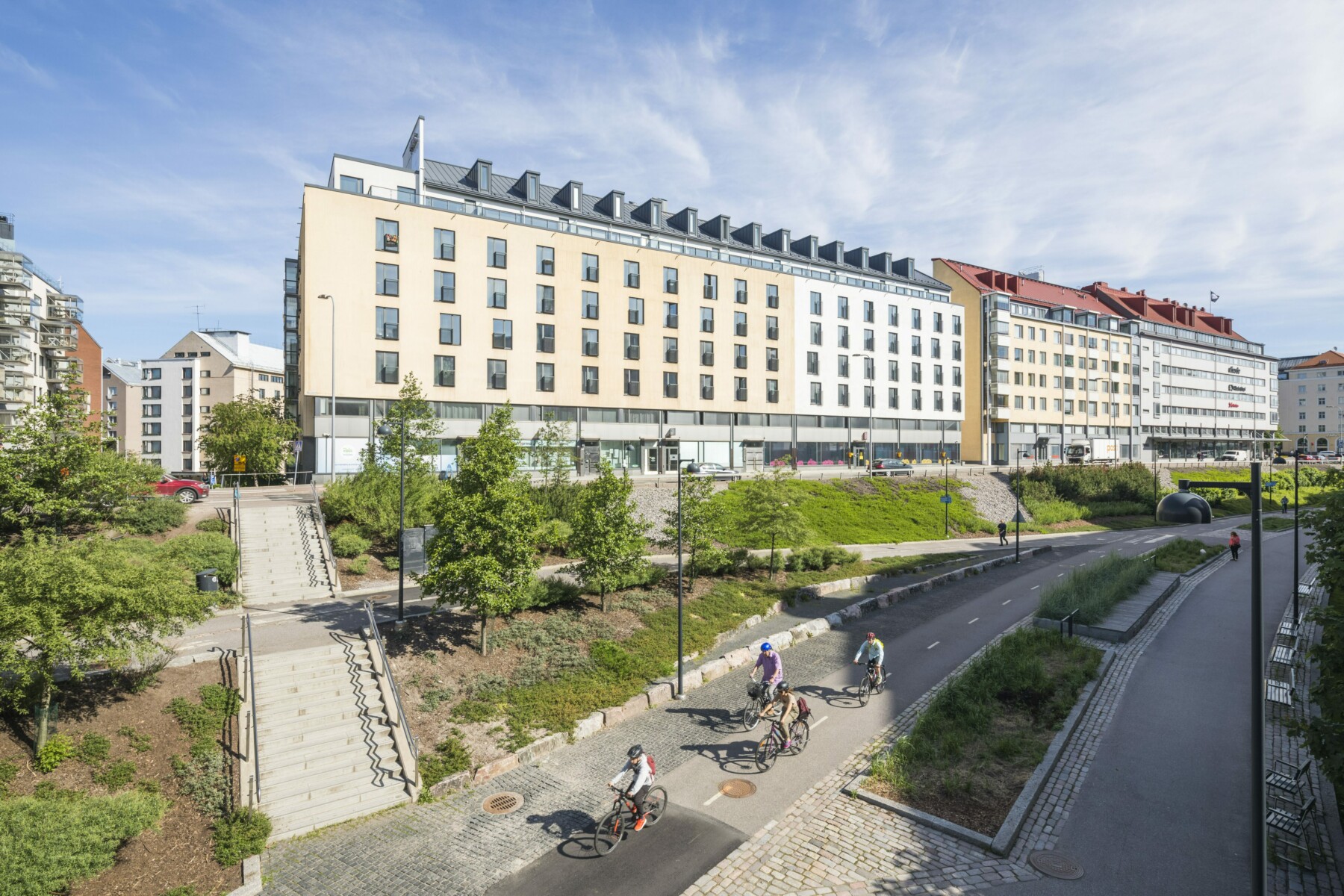 People bike along a paved path beside an embankment and a row of buildings.