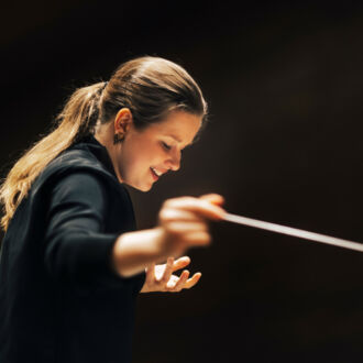 A woman dressed in black gestures with her hands and a conductor’s baton while leading an orchestra.