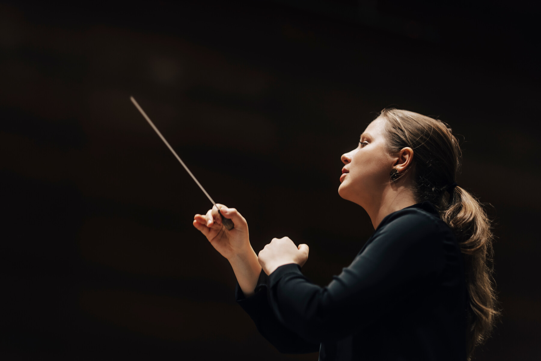 A woman dressed in black gestures with her hands and a conductor’s baton while leading an orchestra.