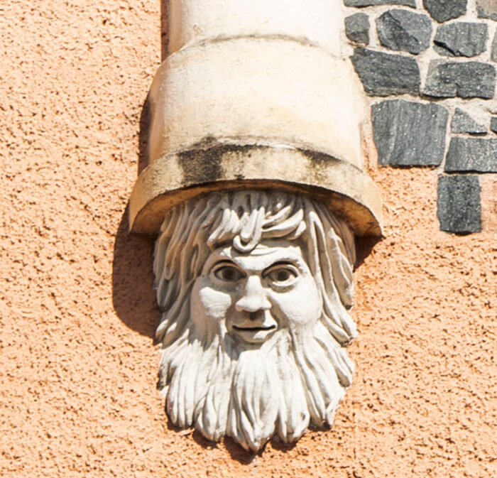 Diese Fotos zeigen eine Nahaufnahme des in Stein gemeißelten gnomartigen Bartgesichts sowie eine größere Ansicht, die deutlich macht, dass das Gesicht ein kleines Detail hoch oben an einem Gebäude zwischen einem Giebel und einem Turm ist.