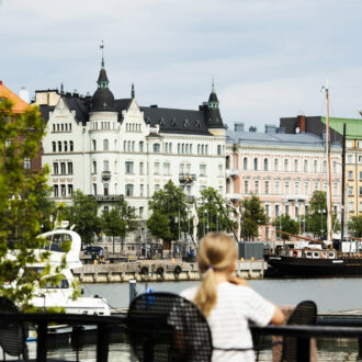 A girl sits at an outdoor café, looking across a bay to a row of ornate buildings on the other side.