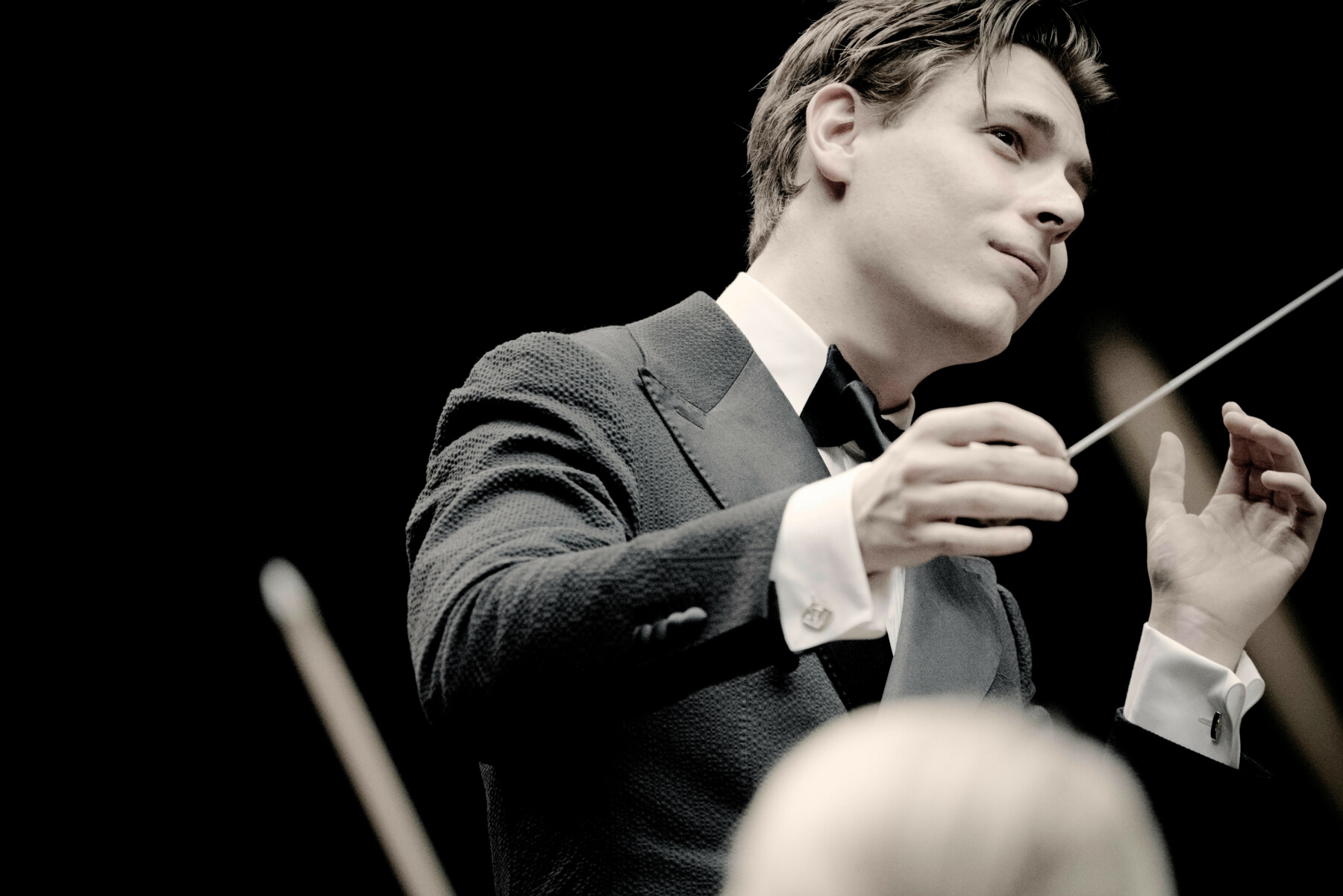 A man dressed in black gestures with his hands and a conductor’s baton while leading an orchestra.