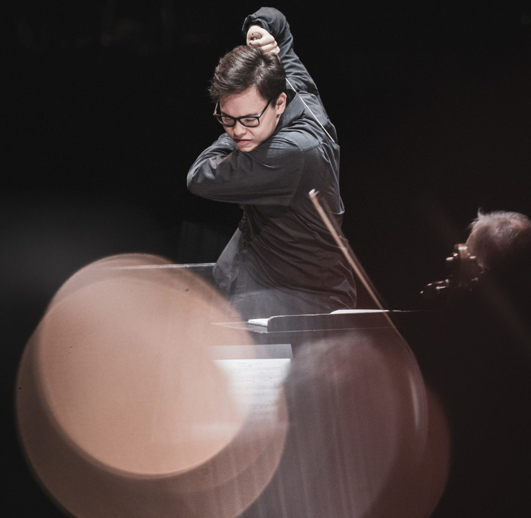 A man dressed in black gestures with his hands and a conductor’s baton while leading an orchestra.