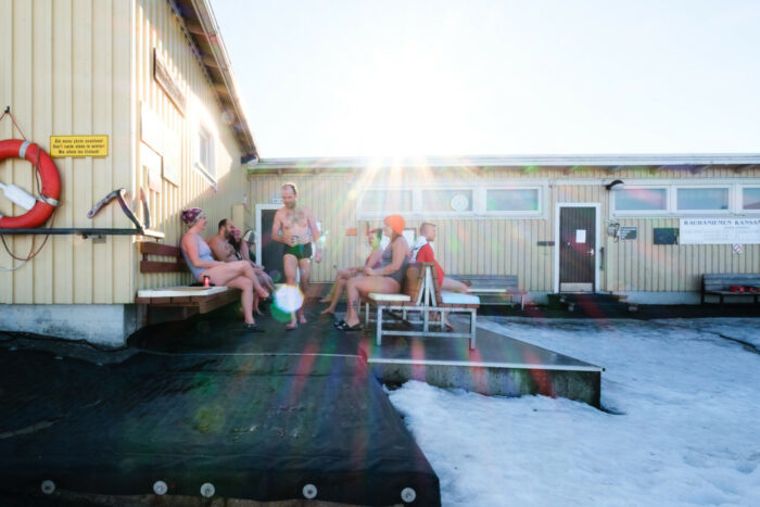 Plusieurs personnes en maillot de bain sont assises sur des bancs dans la cour d’un bâtiment en bois dont le sol est en grande partie enneigé. 