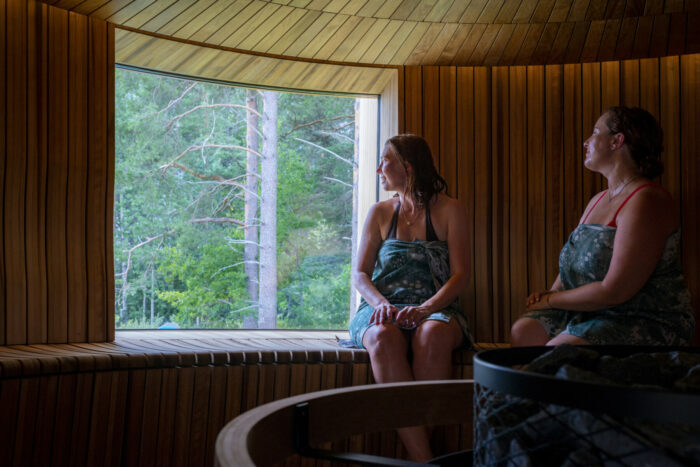 Deux femmes enveloppées dans des serviettes de bain sont assises sur un banc à l’intérieur d’un sauna, le regard tourné vers une fenêtre au-delà de laquelle se profile une forêt.