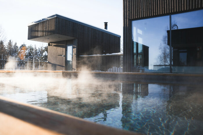 De la vapeur d’eau est en train de s’élever d’une piscine extérieure bordée par des bâtiments en bois aux lignes modernistes.