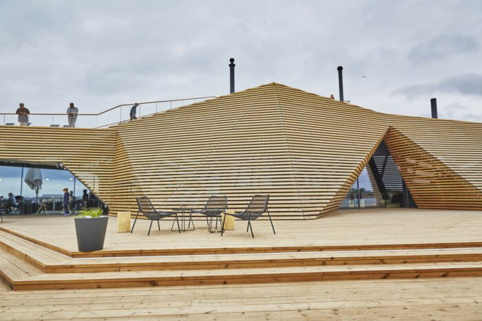 Des chaises sont installées sur un patio devant un bâtiment en bois aux formes anguleuses. 
