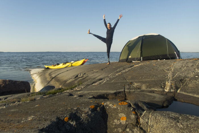 A woman with prosthetic feet is standing on one foot between a tent and two kayaks on a large oceanside rock.