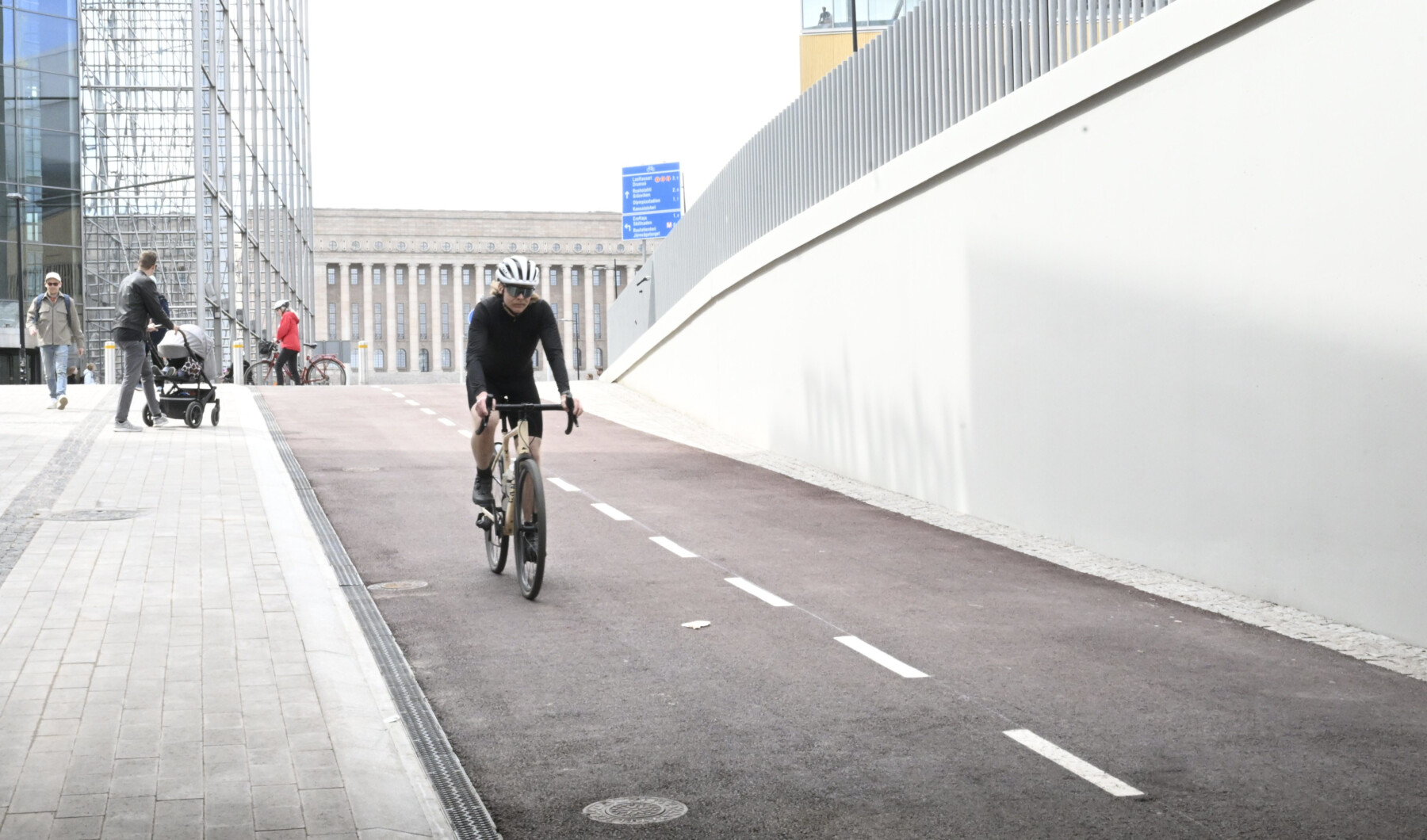 Uma pessoa anda de bicicleta em uma ciclovia pavimentada que desce em direção a um túnel, com prédios e pedestres ao fundo.