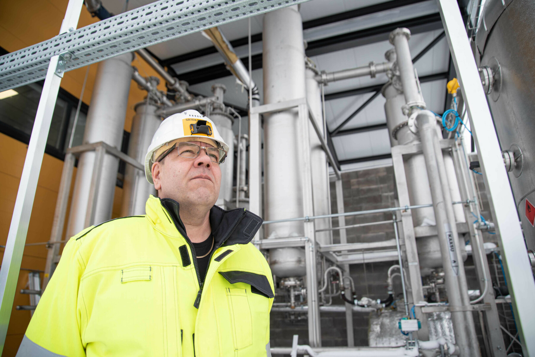 Ein Mann in Arbeitskleidung mit Schutzhelm steht vor einer massiven Struktur aus Metallrohren und Ventilen.