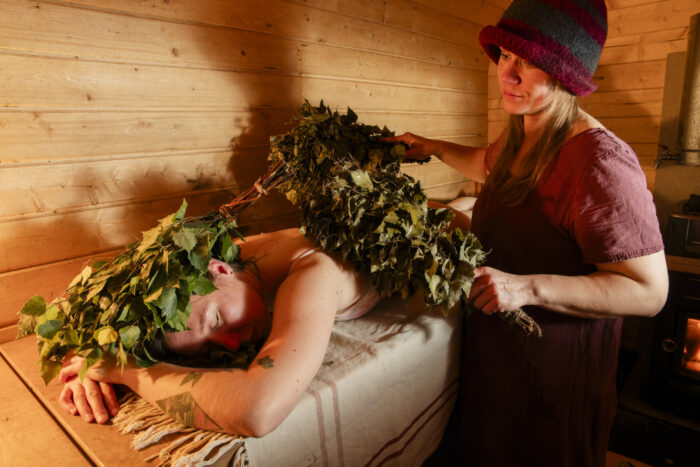 In a wooden room, a person uses a bunch of small tree branches to pat the back of a person lying on a bench.