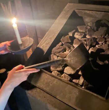 Uma pessoa usa uma concha de cabo longo para despejar água em um suporte de pedras sobre um forno de sauna.