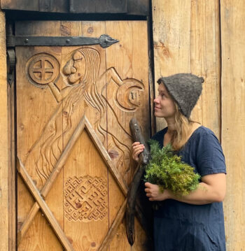 A woman is holding a bunch of leafy branches in front of a wooden door decorated with carvings.