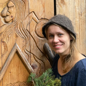 A woman is holding a bunch of leafy branches in front of a wooden door decorated with carvings.