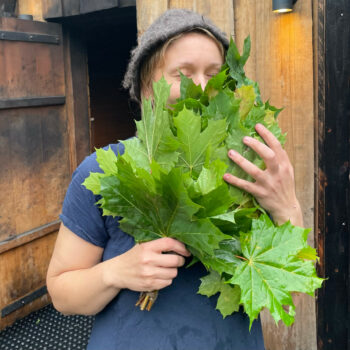 Une femme se tient devant une bâtisse en bois avec dans les mains un bouquet tressé de branchettes d’érable.