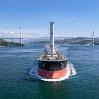 Ein Frachtschiff mit einem ungewöhnlichen zylindrischen Mast segelt direkt auf die Kamera zu, im Hintergrund sind Inseln, eine Brücke und Berge zu sehen.