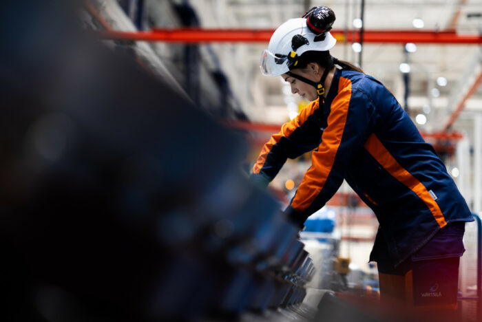 Une femme portant un casque et une combinaison de travail bleue et orange est en train de faire des réglages sur une machine.