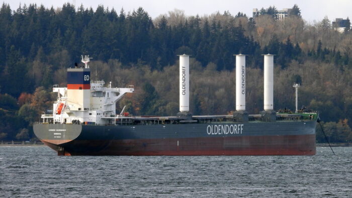 A cargo ship with three unusual cylindrical masts is moored in front of a tree-filled hillside.