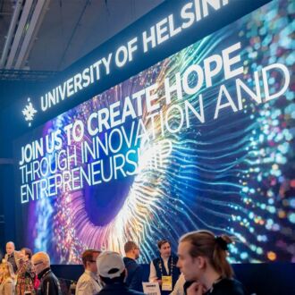 A group of people are talking and looking at screens at the University of Helsinki stand at a startup and tech event.