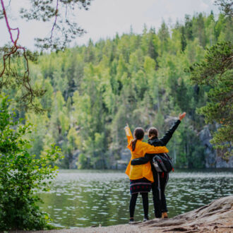 Two people are standing on the shore of a lake in a forest and raising their hands towards the water in a triumphant gesture.