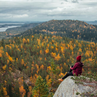 A person sits on a large rock overlooking a forest of green and orange autumn colours and a distant lake.