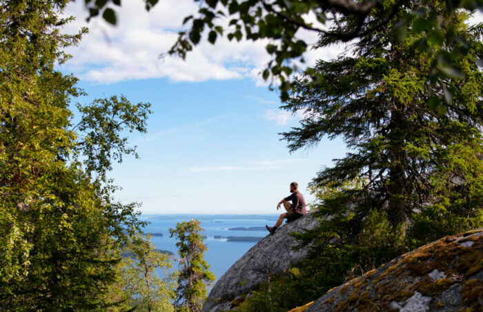 Eine Person sitzt an einem Sommertag auf einem großen Felsen und blickt auf einen See mit Inseln.