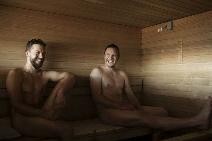 Two mostly naked men are sitting on wooden benches in a wooden room.