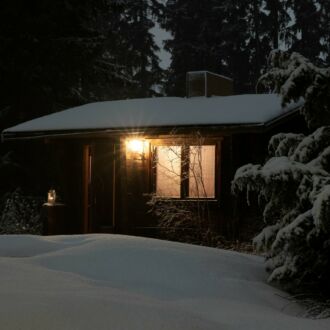 On a dark evening, light shines from the window of a cabin in the middle of snow-covered trees and ground.