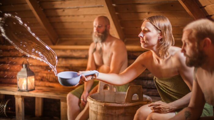 Two men and a woman, all dressed in towels, are sitting in a sauna, and the woman is pouring water from a ladle towards a stove.